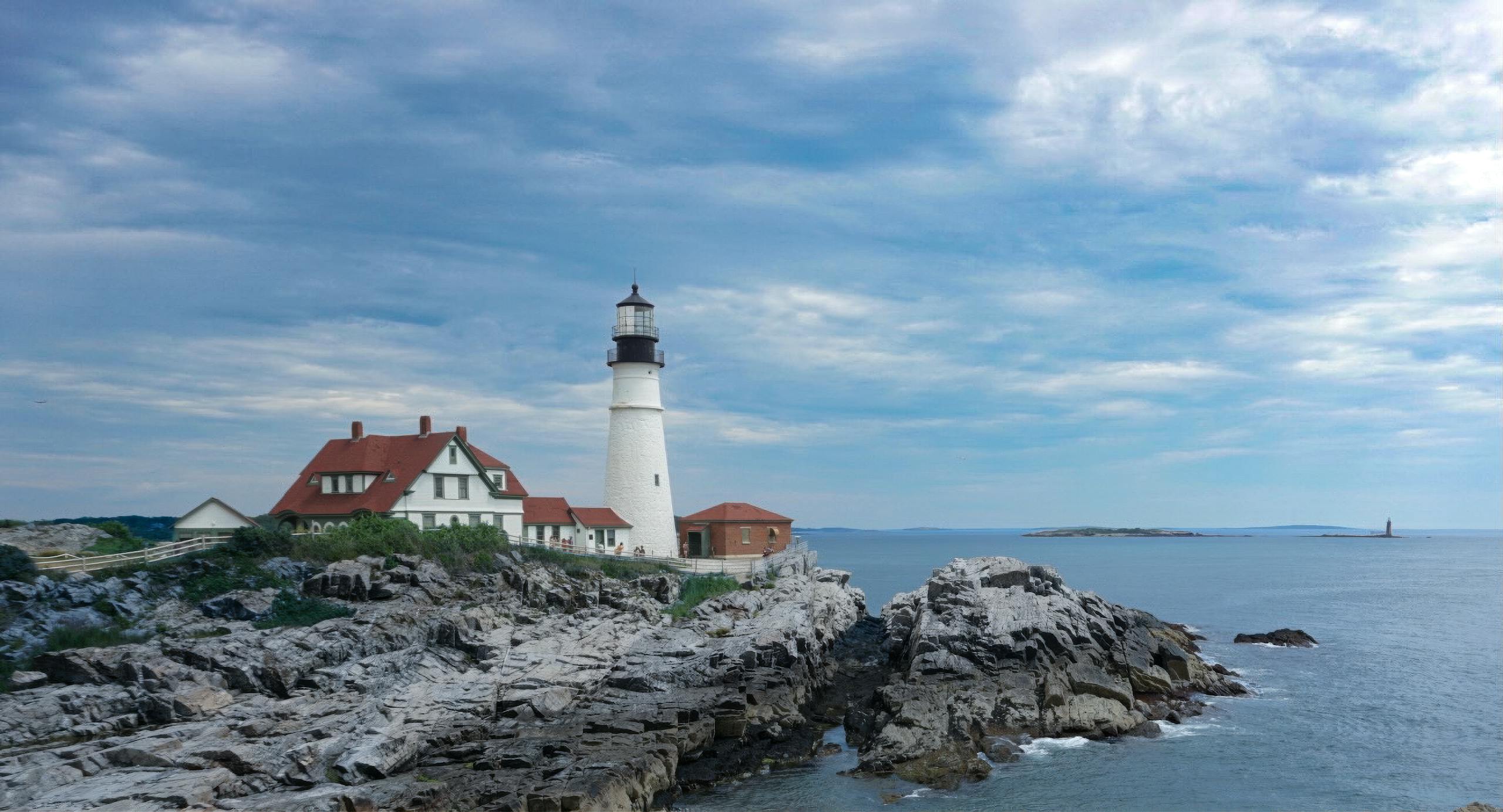 Scenic view of a historic lighthouse perched on rocky cliffs with a vast ocean backdrop and cloudy blue sky.